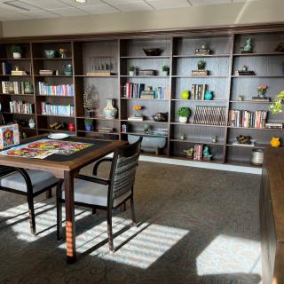 A cozy library space with bookshelves, a table with chairs, and a puzzle in progress, bathed in sunlight.