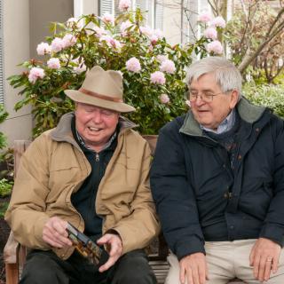Two elderly men sitting on a bench outdoors, surrounded by flowers.