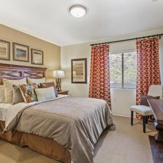A cozy bedroom with a neatly made bed, decorative pillows, and framed artwork on the walls. The room features a lamp on the bedside table and colorful curtains by the window.