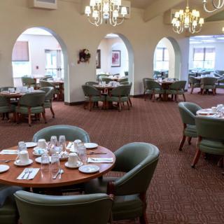 An elegantly decorated dining area featuring multiple round tables set with tableware, surrounded by comfortable chairs, and illuminated by chandeliers.