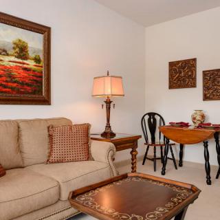 A cozy living room with a sofa, side table with lamp, artwork on the walls, and a small dining area.