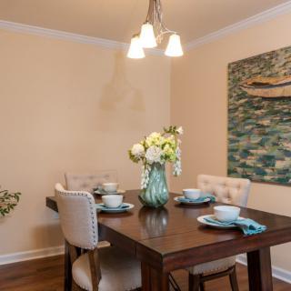 A cozy dining room with a wooden table set for four, featuring elegant place settings and a floral centerpiece. The room is adorned with wall art and illuminated by a modern light fixture.
