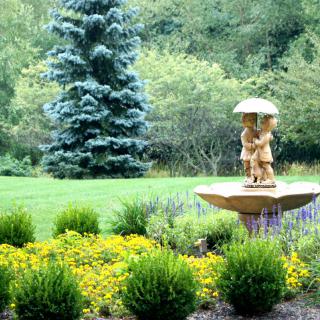 A serene garden landscape featuring a fountain with a sculpture, surrounded by lush greenery.