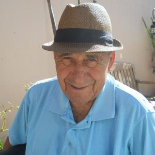 An elderly man wearing a hat and blue shirt, smiling outdoors.