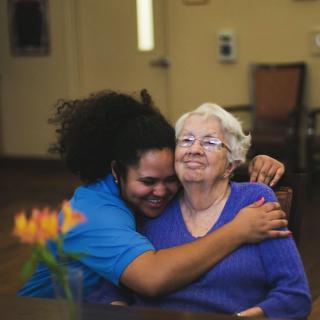 A caregiver warmly hugging an elderly woman, both smiling.