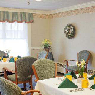 A cozy dining room with neatly arranged tables and chairs, decorated with flowers and a wreath on the wall.