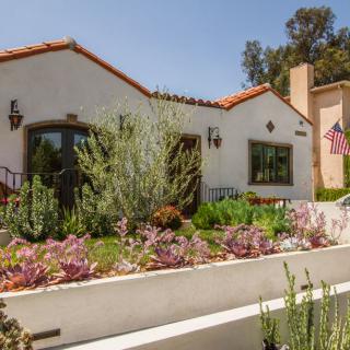 A charming Spanish-style house with a lush succulent garden and an American flag displayed outside on a sunny day.