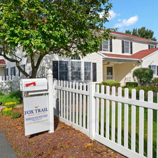 A house with a 'for sale' sign in the front yard, surrounded by a white fence and a tree.