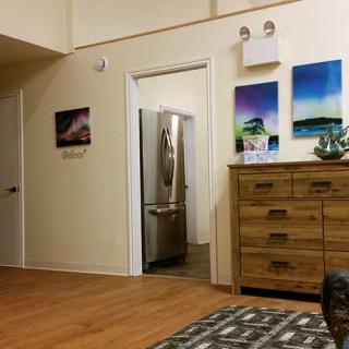 A cozy room with a wooden dresser, colorful paintings on the wall, and a view into the kitchen area.