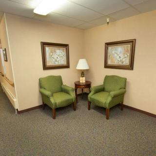 A small waiting area with two green chairs, a lamp on a side table, and framed paintings on the wall.
