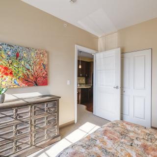 A bright bedroom featuring a colorful painting, a wooden dresser, and an open door revealing a glimpse of another room.