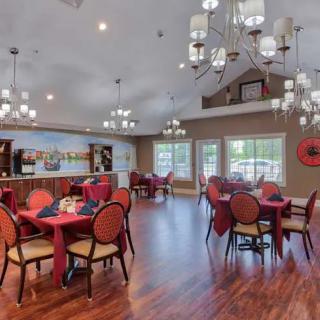 A spacious dining room with elegant chandeliers, multiple tables with red tablecloths, and warm wood flooring.