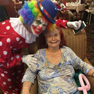 A clown in a colorful costume poses with a smiling elderly woman holding a balloon animal.