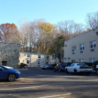 A view of a residential area with two apartment buildings and several parked cars, surrounded by trees in autumn.