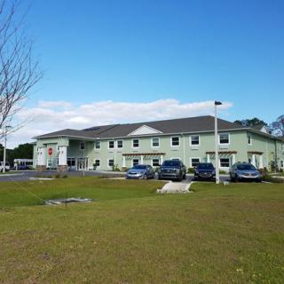 A two-story green building with a parking lot and several cars parked in front, set against a clear blue sky.