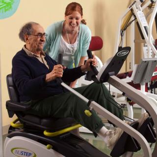 An elderly man working out on gym equipment with assistance from a caregiver.