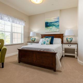 A cozy bedroom featuring a wooden bed, decorative pillows, and matching bedside tables with lamps. A large window allows natural light to fill the room.