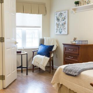 A cozy bedroom with a wooden chair, soft furnishings, and natural light streaming through the window.