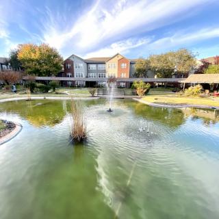 A serene pond with a central fountain and surrounding buildings under a partly cloudy sky.
