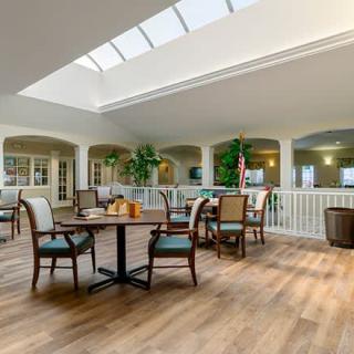 A bright and open dining area with tables and chairs under a skylight, featuring indoor plants and wood flooring.