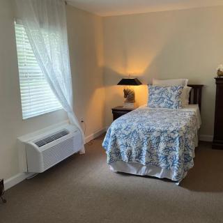 A cozy bedroom featuring a single bed with floral bedding, a bedside table with a lamp, and a window with sheer curtains.