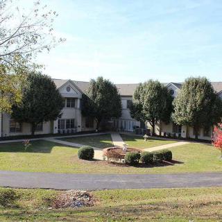 A large mansion surrounded by a well-maintained lawn and trees, with a circular driveway in front.