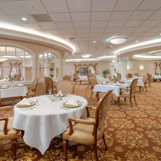 An elegant dining room with neatly arranged tables and chairs, ornate chandeliers, and a carpeted floor.