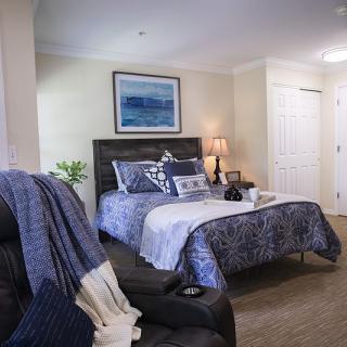 A cozy bedroom with a bed adorned with blue and white bedding, an armchair draped with a throw, and a lamp providing soft lighting, complemented by artwork on the wall.