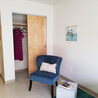 A cozy room with a blue chair near an open closet, featuring a dress and a small table with a book.