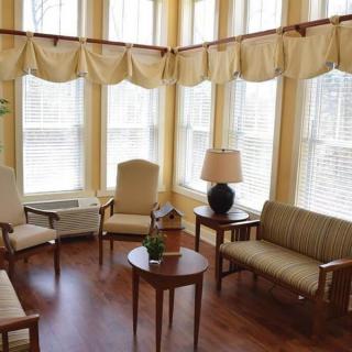 A cozy living room with multiple windows, featuring armchairs, a small sofa, tables, and a lamp.