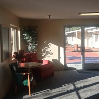 A cozy lobby area with sunlight streaming through the windows, featuring a red armchair, green chair, small table, and potted plant.