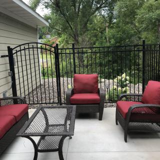 A cozy patio setup with red-cushioned chairs and a metal table, surrounded by a black fence and garden greenery.
