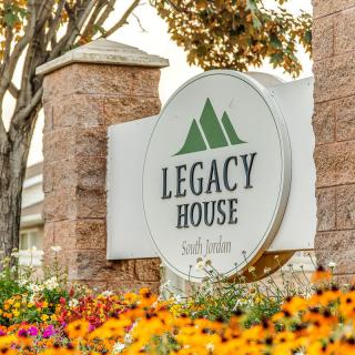 A sign for Legacy House surrounded by colorful flowers and trees.