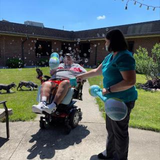 A caregiver playfully blows bubbles for a person in a wheelchair, enjoying a sunny day in the garden.