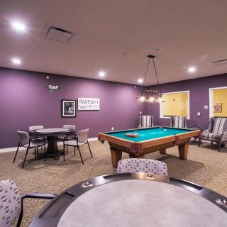 A recreation room featuring a pool table, surrounded by game tables and chairs, with purple walls and ambient indoor lighting.