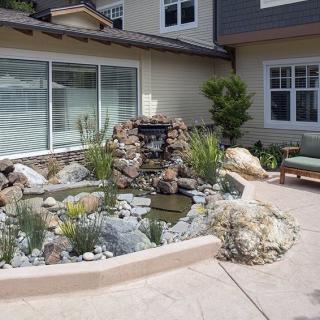 A beautifully landscaped patio featuring a small pond with a fountain, surrounded by rocks and greenery, adjacent to a house.