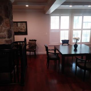 A dining area with a wooden table and chairs, featuring large windows and hardwood flooring.