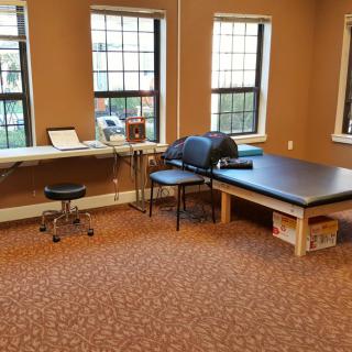 A therapy treatment room with a table, chairs, and medical equipment against the backdrop of large windows.