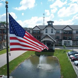 An American flag waves in front of a large building with a pond and fountain, set against a cloudy sky.