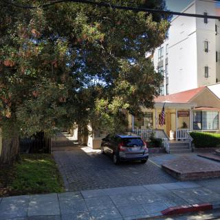 A suburban house with a driveway and parked car, surrounded by trees, adjacent to a tall building.