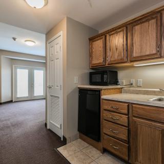 A view of a small kitchenette with wooden cabinets and a microwave, leading to a living area with large doors.