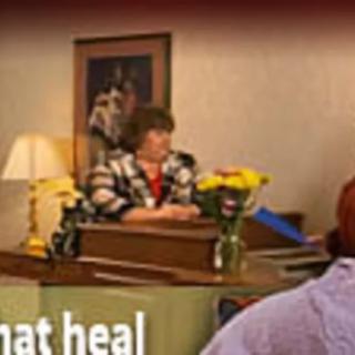 A woman sits at a desk in an office decorated with flowers and a lamp.