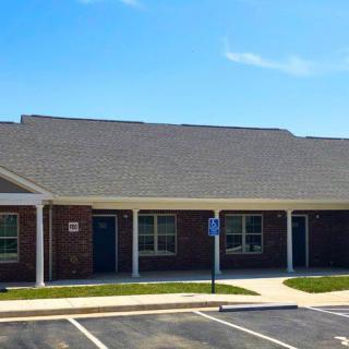 A single-story brick residential building with a sloped roof and an adjacent parking lot under a clear blue sky.