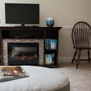 A cozy living room with a TV stand and electric fireplace, featuring a nearby chair and neatly arranged books on a shelf.