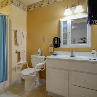 A cozy bathroom with a yellow accent wall, featuring a white vanity and sink, toilet with support bars, mirror with overhead lighting, and a shower with a blue curtain.