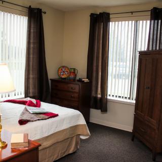 A cozy bedroom with natural light shining through the windows, featuring a neatly made bed with pillows and a bedside lamp.