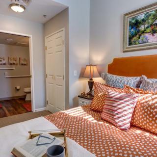 A cozy bedroom featuring a neatly made bed with patterned pillows, a side lamp, and artwork on the wall.