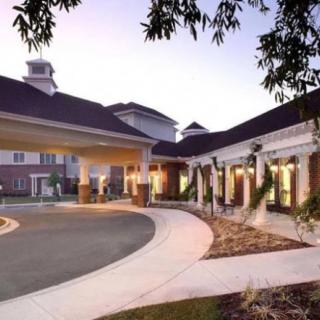A well-lit building entrance with a driveway, surrounded by landscaped gardens, at dusk.