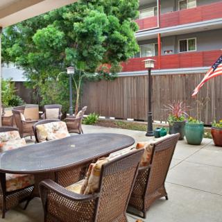A cozy patio with wicker furniture and a dining table, surrounded by greenery and an American flag in the background.