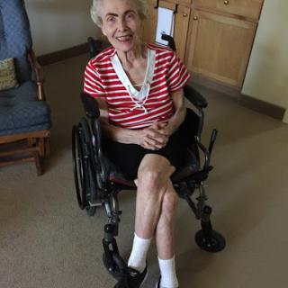 An elderly woman sitting in a wheelchair, wearing a striped shirt and smiling warmly.